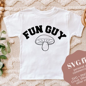 Pode incluir: Camiseta branca com texto preto que diz "FUN GUY" e um gráfico de cogumelo preto e branco. O texto está em uma fonte universitária. A camiseta está sobre uma superfície marrom claro com uma cesta de vime e um galho de vegetação.