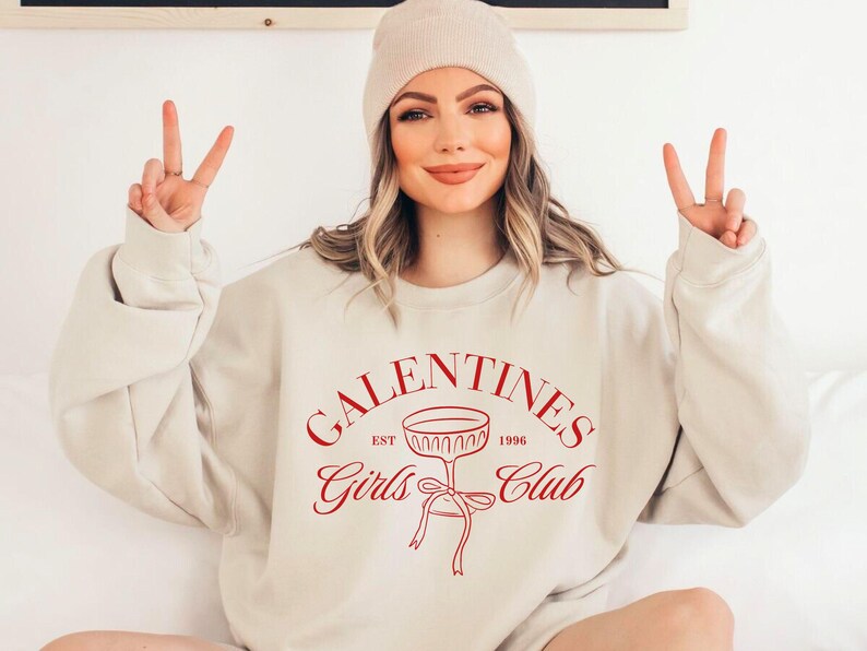 May include: A beige sweatshirt with the text "Galentines Girls Club Est. 1996" in red lettering. The design also includes a red illustration of a champagne glass with a bow.