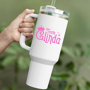 Pu&ograve; includere: Tazza da viaggio bianca con una corona rosa e il testo "Team Glinda" con stelle glitter rosa.