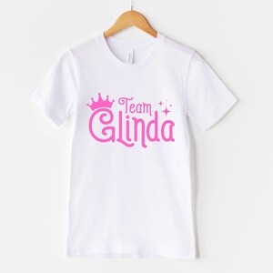 Pu&ograve; includere: Maglietta bianca con una corona rosa, stelle e il testo "Team Glinda" stampato sul davanti.