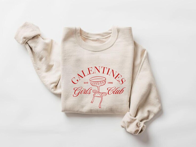 May include: A beige sweatshirt with the text "Galentines Girls Club Est. 1996" in red lettering and a red outline of a champagne glass with a bow.
