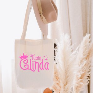 Pu&ograve; includere: Una borsa tote in tela bianca con testo rosa che recita "Team Glinda" con una corona rosa sopra il testo. La borsa &egrave; appesa a una parete bianca con una pianta bianca e soffice sullo sfondo.