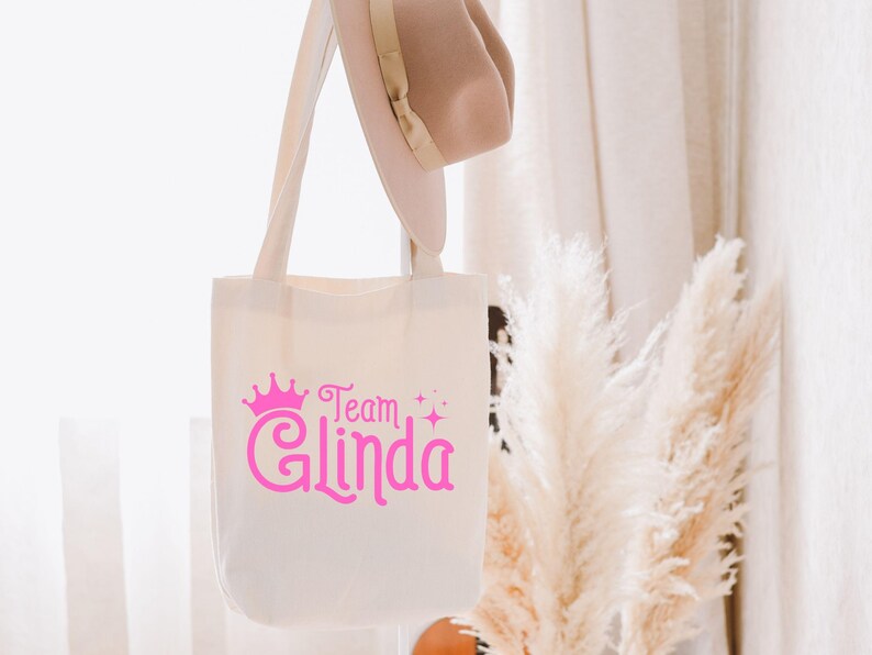 Pu&ograve; includere: Una borsa tote in tela bianca con testo rosa che recita "Team Glinda" con una corona rosa sopra il testo. La borsa &egrave; appesa a una parete bianca con una pianta bianca e soffice sullo sfondo.