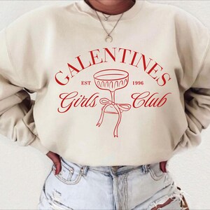 May include: A cream-colored sweatshirt with the text "Galentines Girls Club" in red lettering. The text is in a circle with a champagne glass graphic in the center. The text "Est 1996" is above the glass and the text "Girls Club" is below the glass.