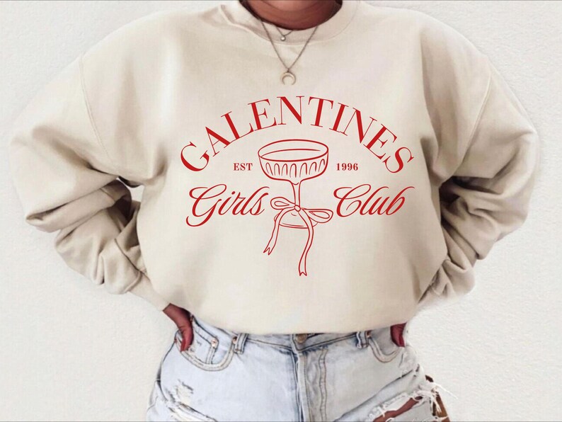May include: A cream-colored sweatshirt with the text "Galentines Girls Club" in red lettering. The text is in a circle with a champagne glass graphic in the center. The text "Est 1996" is above the glass and the text "Girls Club" is below the glass.