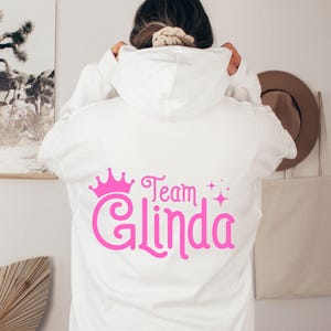 Pu&ograve; includere: Felpa bianca con una corona rosa e il testo "Team Glinda" sul retro.