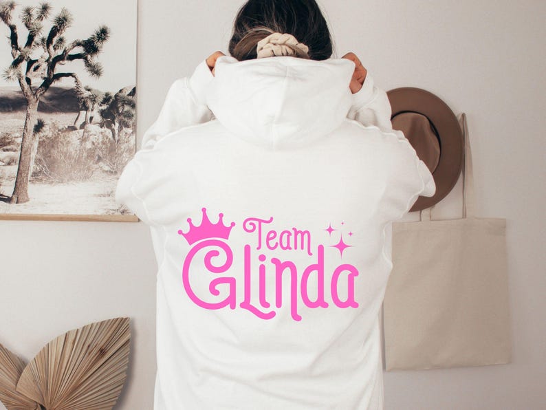 Pu&ograve; includere: Felpa bianca con una corona rosa e il testo "Team Glinda" sul retro.