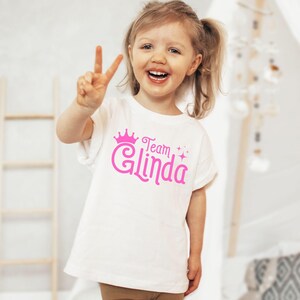 Pu&ograve; includere: Una maglietta bianca con una corona rosa e il testo "Team Glinda" in lettere rosa.