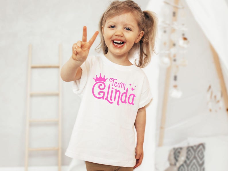Pu&ograve; includere: Una maglietta bianca con una corona rosa e il testo "Team Glinda" in lettere rosa.