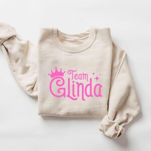Pu&ograve; includere: Una felpa beige con una corona rosa e il testo "Team Glinda" stampato sul davanti.