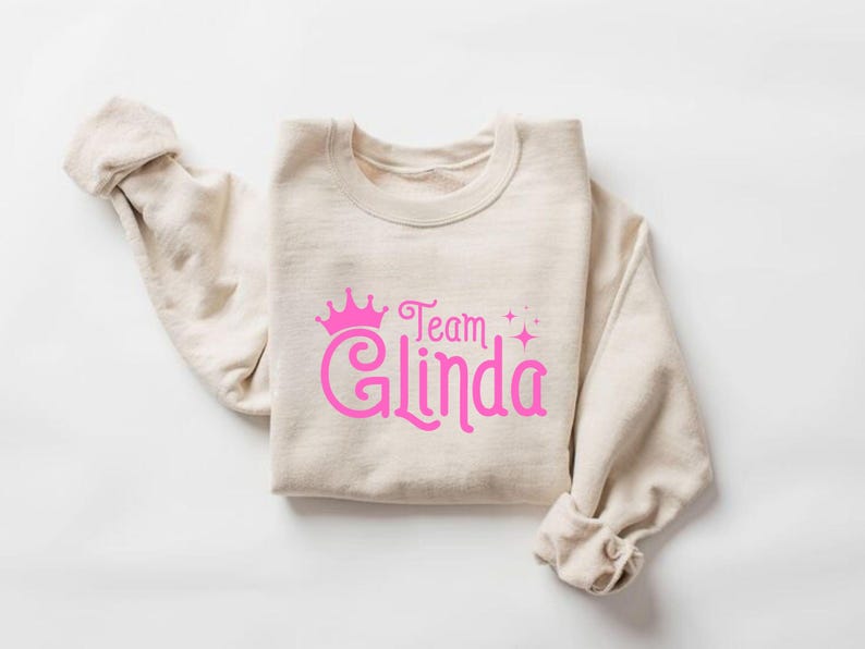 Pu&ograve; includere: Una felpa beige con una corona rosa e il testo "Team Glinda" stampato sul davanti.