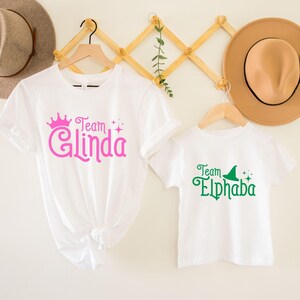 Pu&ograve; includere: Due magliette bianche con un design grafico verde e rosa. La maglietta pi&ugrave; grande dice "Team Glinda" con una corona rosa e glitter. La maglietta pi&ugrave; piccola dice "Team Elphaba" con un cappello da strega verde e glitter.