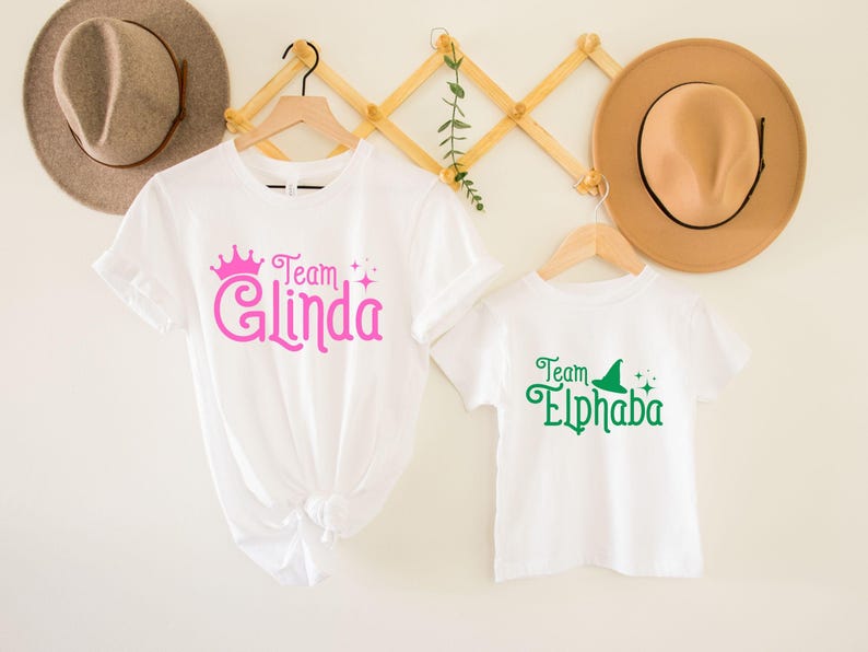 Pu&ograve; includere: Due magliette bianche con un design grafico verde e rosa. La maglietta pi&ugrave; grande dice "Team Glinda" con una corona rosa e glitter. La maglietta pi&ugrave; piccola dice "Team Elphaba" con un cappello da strega verde e glitter.