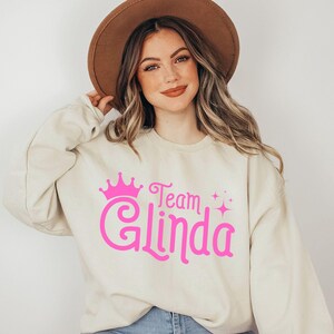 Pu&ograve; includere: Una felpa color crema con una corona rosa e il testo "Team Glinda" in rosa, in un font stilizzato.