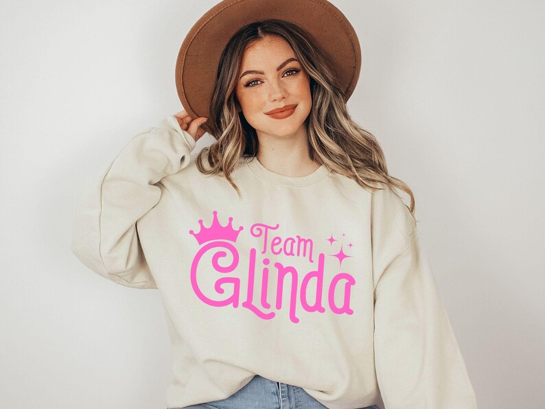 Pu&ograve; includere: Una felpa color crema con una corona rosa e il testo "Team Glinda" in rosa, in un font stilizzato.