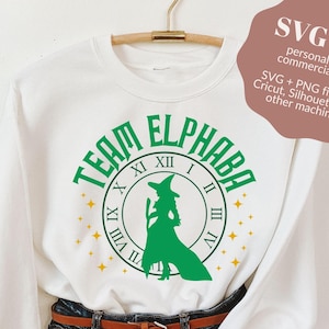 May include: White sweatshirt with a green and gold design that says "Team Elphaba" with a witch silhouette inside a clock face.
