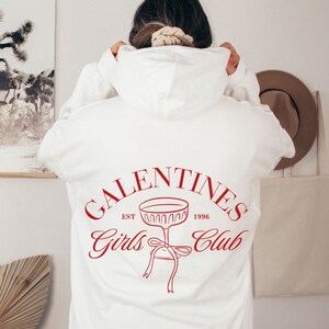 May include: White hoodie with a red graphic print that says "Galentines Girls Club Est 1996" with a stylized image of a champagne glass with a bow.