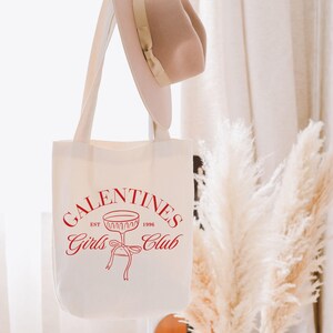 May include: White canvas tote bag with red text that reads "Galentines Girls Club Est. 1998" and a red outline of a champagne glass with a ribbon.