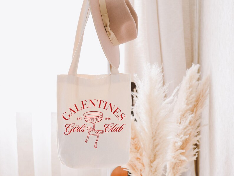 May include: White canvas tote bag with red text that reads "Galentines Girls Club Est. 1998" and a red outline of a champagne glass with a ribbon.