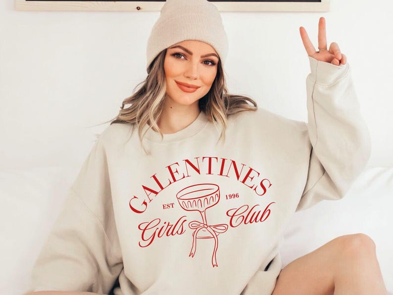 May include: A beige sweatshirt with the text "Galentines Girls Club Est 1996" in red lettering. The sweatshirt features a red illustration of a champagne glass with a bow.