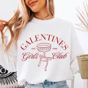 May include: White sweatshirt with red text that reads "Galentines Girls Club Est. 1996" with a red illustration of a champagne glass with a bow.
