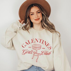 May include: A cream-colored sweatshirt with a red graphic that says "Galentines Girls Club Est. 1996" with a wine glass illustration.