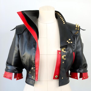 Może przedstawiać: Czarna kurtka ze sztucznej skóry z czerwonymi akcentami, prezentowana na manekinie. Kurtka posiada szeroki kołnierz, złote okucia i detale z ćwiekami na ramionach i mankietach. Krótka kurtka ma odważny, odważny design.