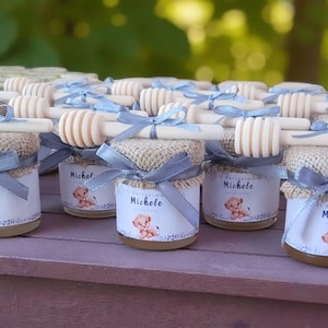 May include: Small glass jars filled with honey, each topped with a wooden honey dipper and burlap fabric. The jars are adorned with light blue ribbons and labels featuring a cartoon lion and the name "Michele".