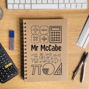 May include: A brown notebook with a spiral binding. The cover features a black and white illustration of a calculator, plus and minus signs, a division sign, a multiplication sign, and the text "Mr McCabe". The notebook also has a black and white illustration of a pie chart, a protractor, and mathematical equations.