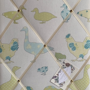 May include: A white fabric notice board with a grid pattern of white ribbon. The board is decorated with a repeating pattern of yellow and blue ducks and chickens. The board has gold push pins and a small white card with text that reads "Rosie Posie Handmade Designs,  Luxury Fabric,  Gift Wrap,  Cards,  Linen Cushions,  UK"