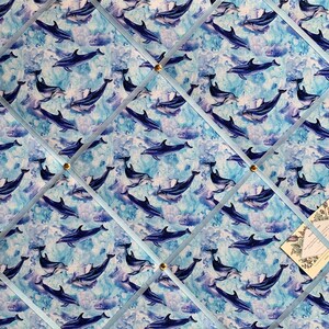May include: A blue and white fabric bulletin board with a repeating pattern of dolphins swimming in the ocean. The board is divided into sections by blue ribbon and has gold push pins.