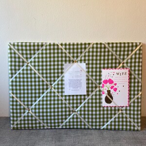 May include: A rectangular memo board covered in green and white gingham fabric. Cream-colored ribbons crisscross the surface, secured by gold-colored tacks. A card with pink flowers and the word "WIFE" is pinned to the board, along with a white paper.