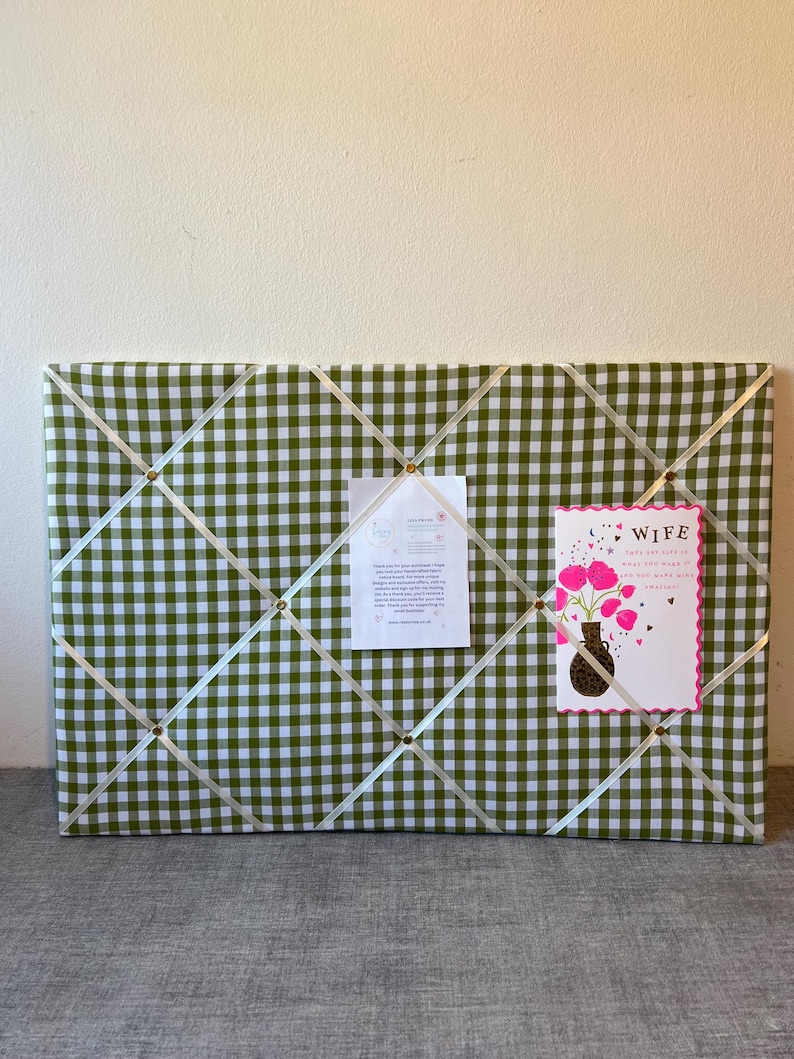 May include: A rectangular memo board covered in green and white gingham fabric. Cream-colored ribbons crisscross the surface, secured by gold-colored tacks. A card with pink flowers and the word "WIFE" is pinned to the board, along with a white paper.