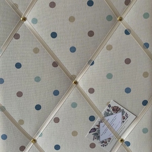May include: A white fabric notice board with a polka dot pattern in blue, brown, and beige. The board has white ribbon crisscrossing the surface and gold tacks holding the ribbon in place. A business card is pinned to the board.