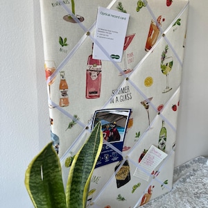 May include: A white cork board with a summer cocktail theme. The board is covered in a white fabric with a pattern of cocktails, fruit, and text. The board has white ribbon crisscrossing it and gold push pins holding up various items, including a business card, a photo, and a handwritten shopping list. The text on the board reads "Summer in a glass", "Pink Lady", "Mint", "Lowess Gin", and "Nisbets Gin".