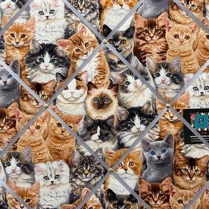 May include: A corkboard covered in a fabric featuring a repeating pattern of adorable kittens with different fur colours and patterns. The kittens are arranged in a diamond pattern and are looking at the viewer.