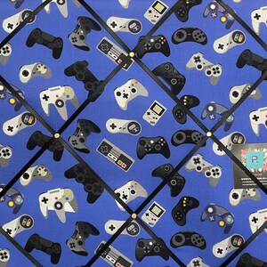 May include: A blue fabric notice board with black ribbon crisscrossing the surface. The board is covered in a pattern of various video game controllers in black, white, and grey. The controllers are arranged in a diagonal pattern.