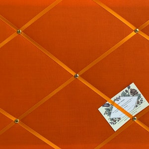 以下が含まれることがあります： オレンジ色の布製の掲示板で、金色のリボンが表面を交差しています。金色の画鋲がリボンを固定しています。小さなカードにテキストが書かれていて、掲示板にピン止めされています。