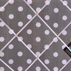 May include: A gray fabric bulletin board with white polka dots and white ribbon crisscrossing the board. Gold tacks are used to hold the ribbon in place. A business card is pinned to the board.