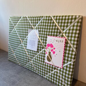 May include: A green and white gingham fabric memo board with white ribbon crisscrossing the surface. Two cards are pinned to the board. One card says "WIFE" and features a vase of pink flowers. The other card has text.