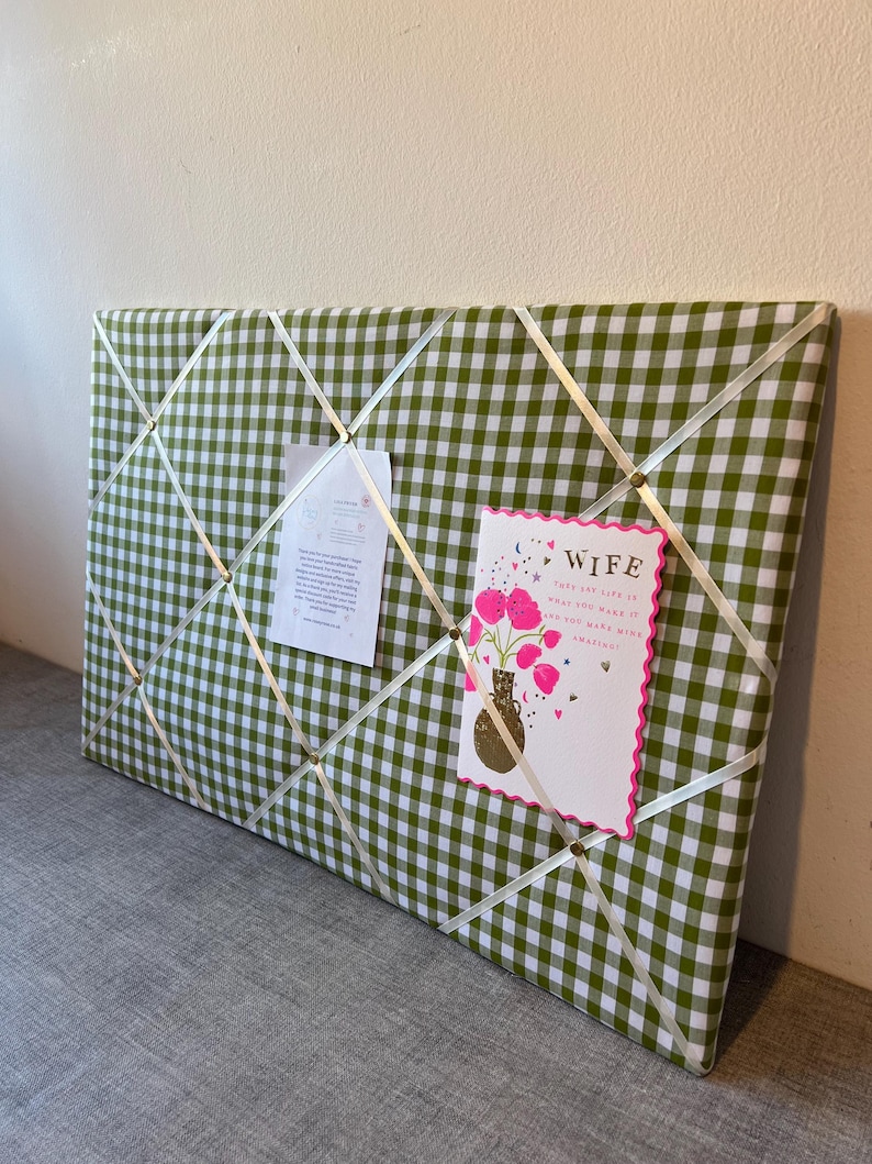 May include: A green and white gingham fabric memo board with white ribbon crisscrossing the surface. Two cards are pinned to the board. One card says "WIFE" and features a vase of pink flowers. The other card has text.