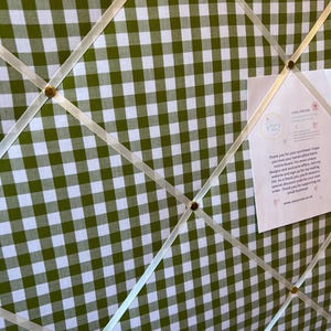 May include: A green and white gingham fabric memo board with cream-colored ribbons and gold-colored tacks. A small card is attached to the board with text. The board is designed for displaying notes and photos.