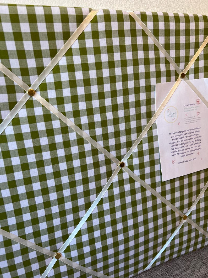 May include: A green and white gingham fabric memo board with cream-colored ribbons and gold-colored tacks. A small card is attached to the board with text. The board is designed for displaying notes and photos.