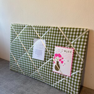 May include: A rectangular memo board covered in green and white gingham fabric. White ribbons crisscross the surface, holding a card that says "Wife" and a small paper. The card features pink flowers in a vase.