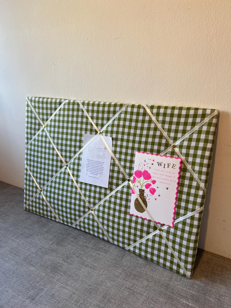 May include: A rectangular memo board covered in green and white gingham fabric. White ribbons crisscross the surface, holding a card that says "Wife" and a small paper. The card features pink flowers in a vase.