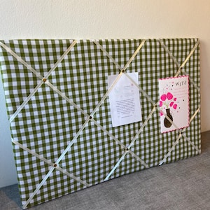 May include: A green and white gingham fabric memo board with cream-colored ribbon crisscrossing the surface. Small gold-colored tacks secure the ribbon. Two cards are pinned to the board. One card says "Wife" and the other has text.