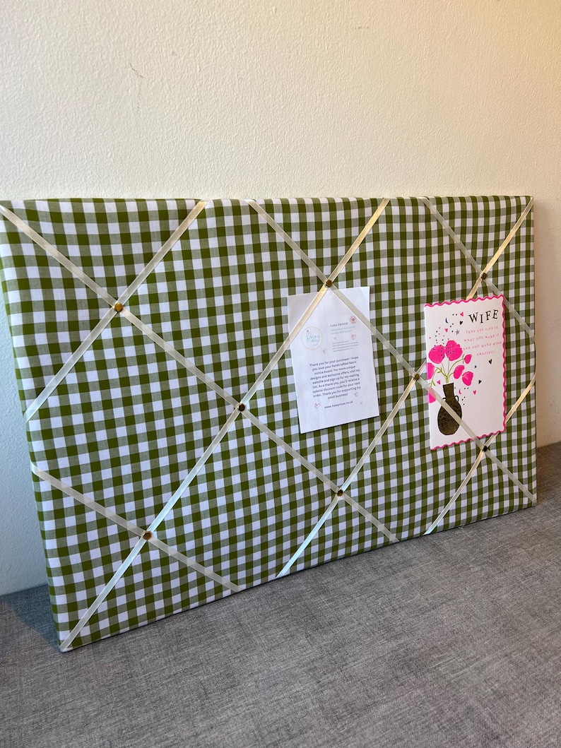 May include: A green and white gingham fabric memo board with cream-colored ribbon crisscrossing the surface. Small gold-colored tacks secure the ribbon. Two cards are pinned to the board. One card says "Wife" and the other has text.