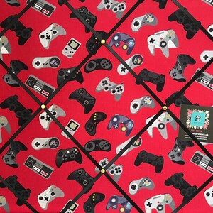 May include: A red fabric notice board with black ribbon crisscrossing the surface. The board is covered in a repeating pattern of video game controllers in various colours and styles. The controllers are arranged in a diamond pattern.