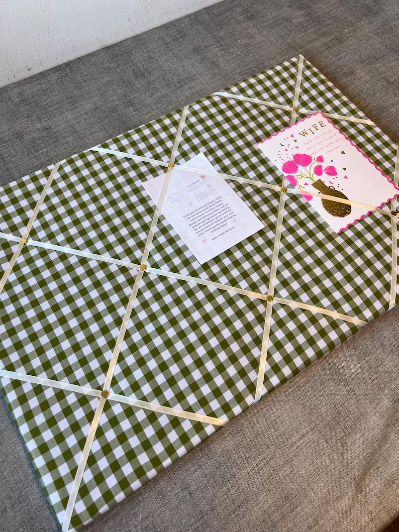 May include: A green and white gingham fabric memo board with cream-colored ribbons and gold-colored tacks. A greeting card that says "Wife" and a small white card are attached to the board.