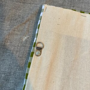 May include: Close-up of the back of a canvas art piece. The canvas is a natural beige color, with a green and white gingham pattern along the edges. A metal D-ring is attached for hanging.